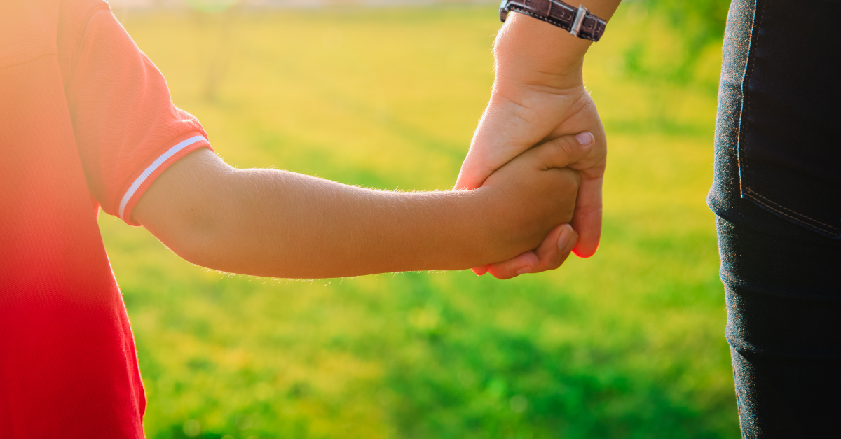 A child holding the hand of an adult walking in the sunshine.