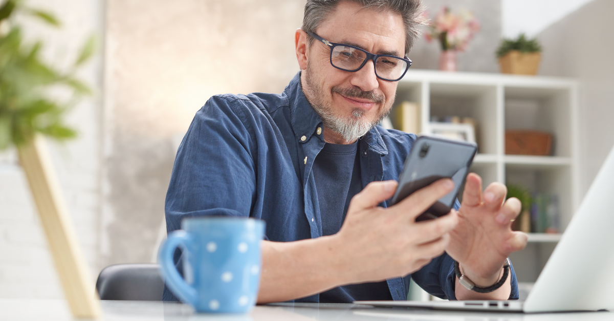 A man smiling and looking at his smartphone with his laptop in the foreground